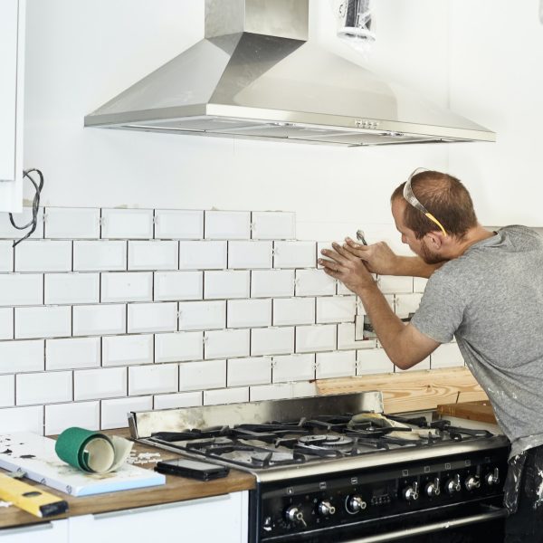 A man working in a new kitchen, a tiler applying tiles to the wall behind the cooker.